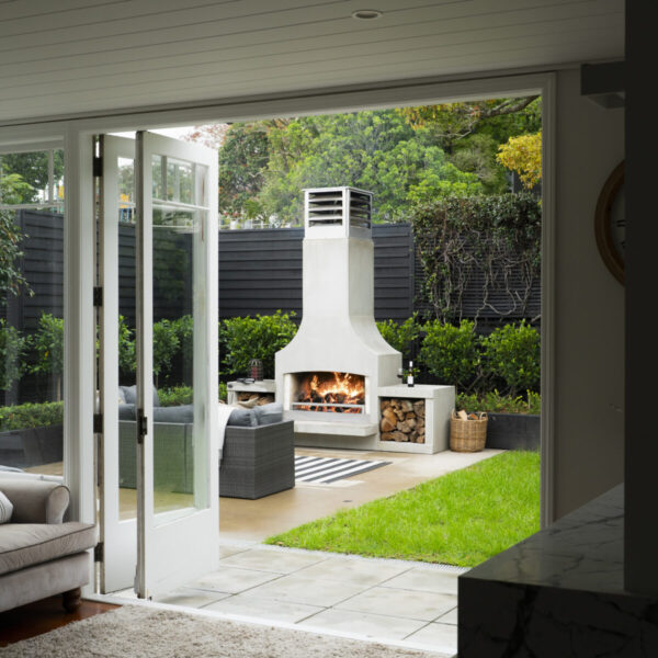Outdoor fireplace lit on the lawn, viewed through open bifold doors from inside the house