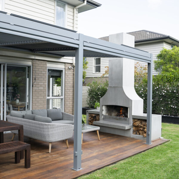 White outdoor fireplace under a louvred pergola, proportioned to a small deck and lawn