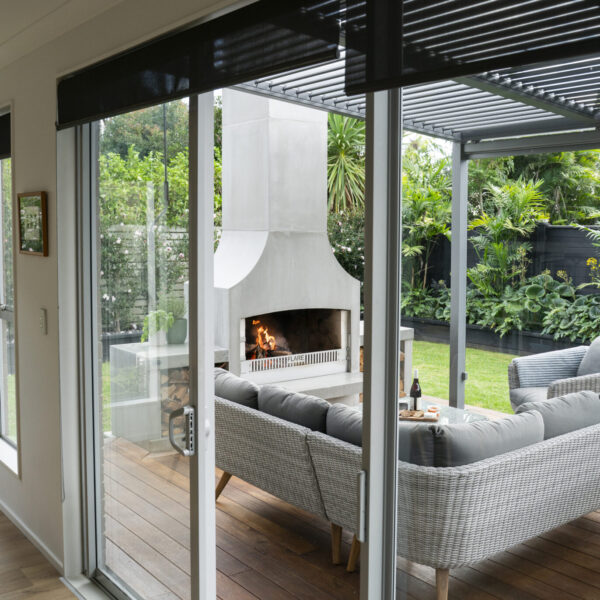 White outdoor fireplace framed by glass doors, with a louvred roof and wicker seating beyond