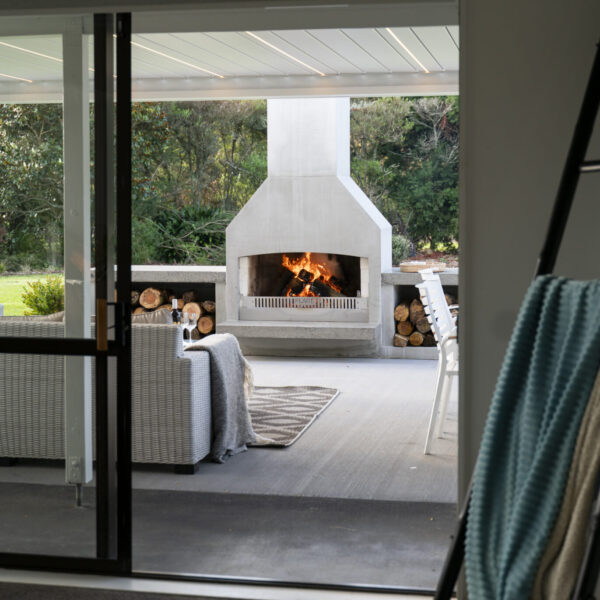 Fire burning in an outdoor fireplace, visible from an indoor living space through wide glass doors
