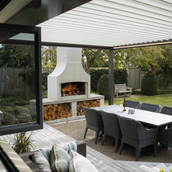 Outdoor fireplace and dining table sheltered under a louvred pergola on a paved patio