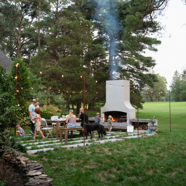 White outdoor fireplace on a lawn with tall pines behind and a group gathered at a table